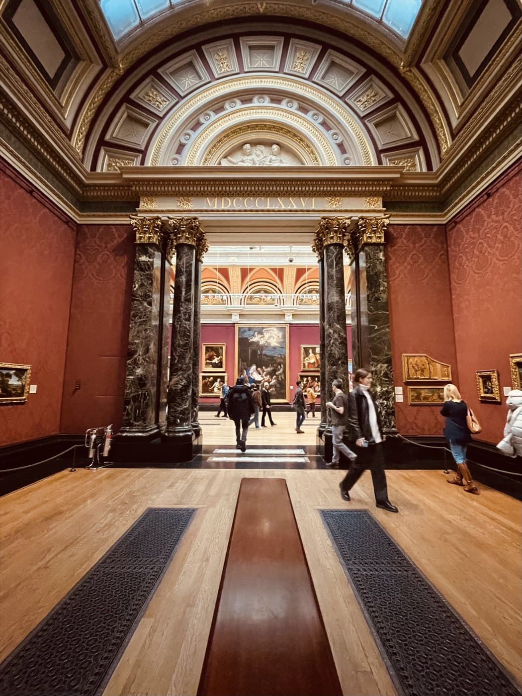 A grand red-walled gallery hall with marble columns and a gilded arch