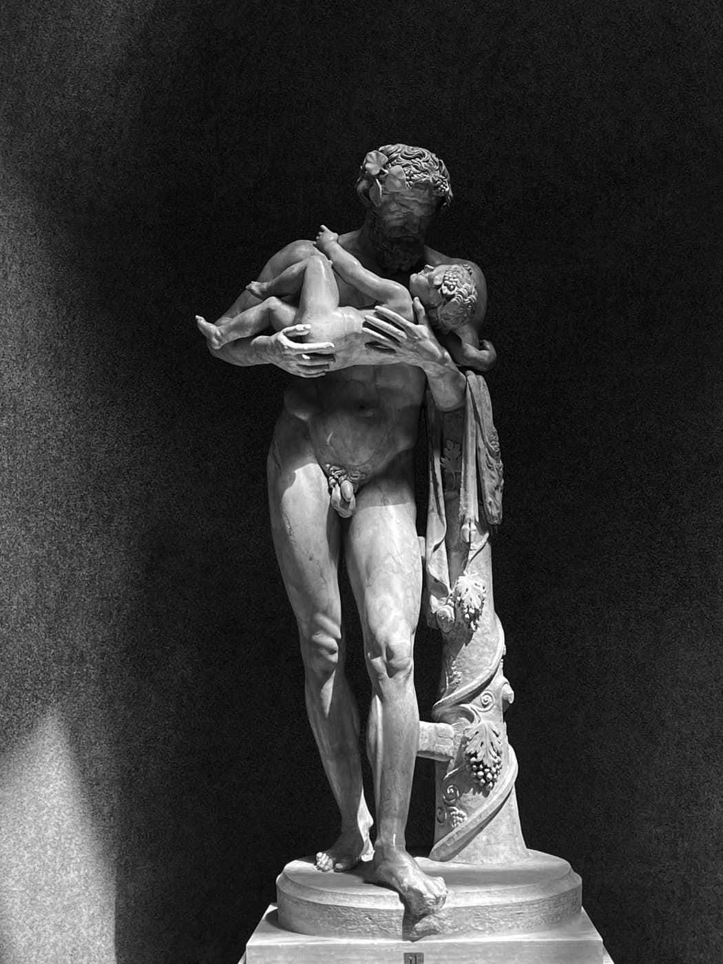 A marble statue of a bearded man carrying a child, lit against a dark museum wall